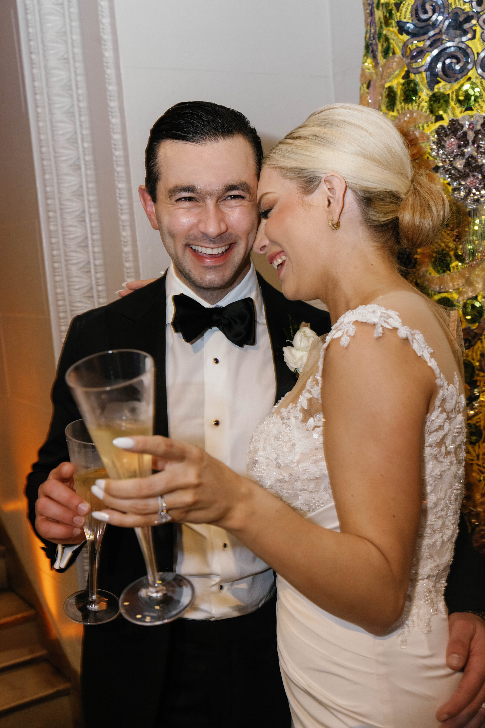 Bride and groom laugh together holding two champagne flutes beside a beaded sequinned costume.